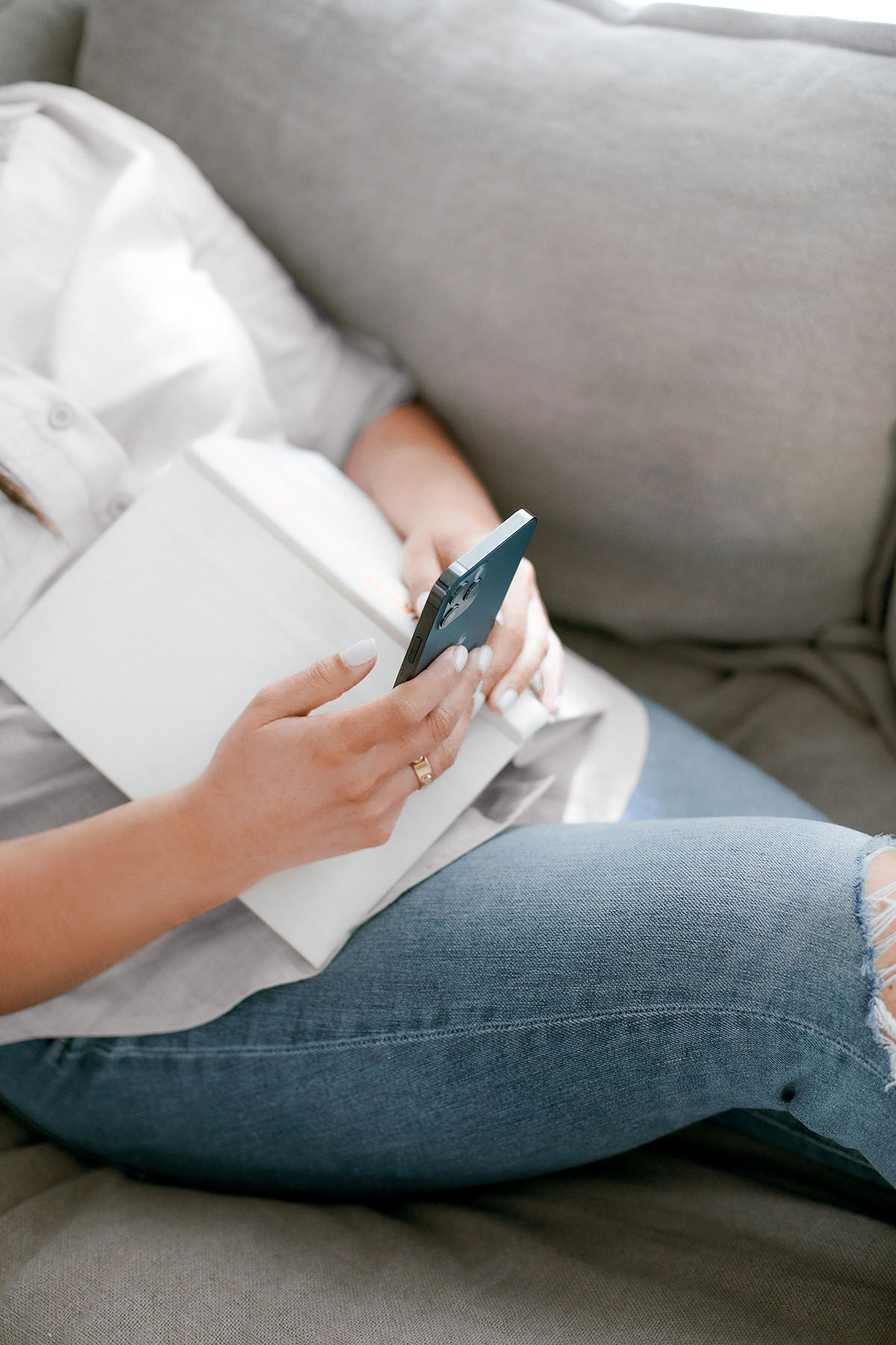 Person sitting on a couch, holding a smartphone and a book in hand, wearing a white shirt and ripped jeans.