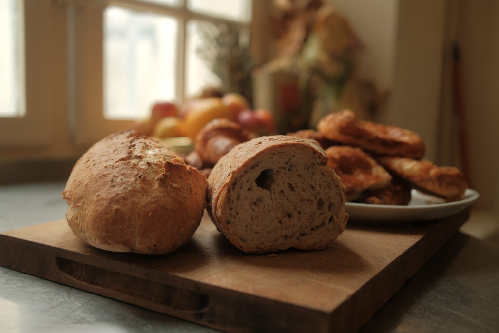 A sliced loaf of bread rests on a wooden board, inviting you to savor each piece. In the background, a plate of pastries and a bowl of fruit reflect a delightful non-diet approach to enjoying life’s simple pleasures.