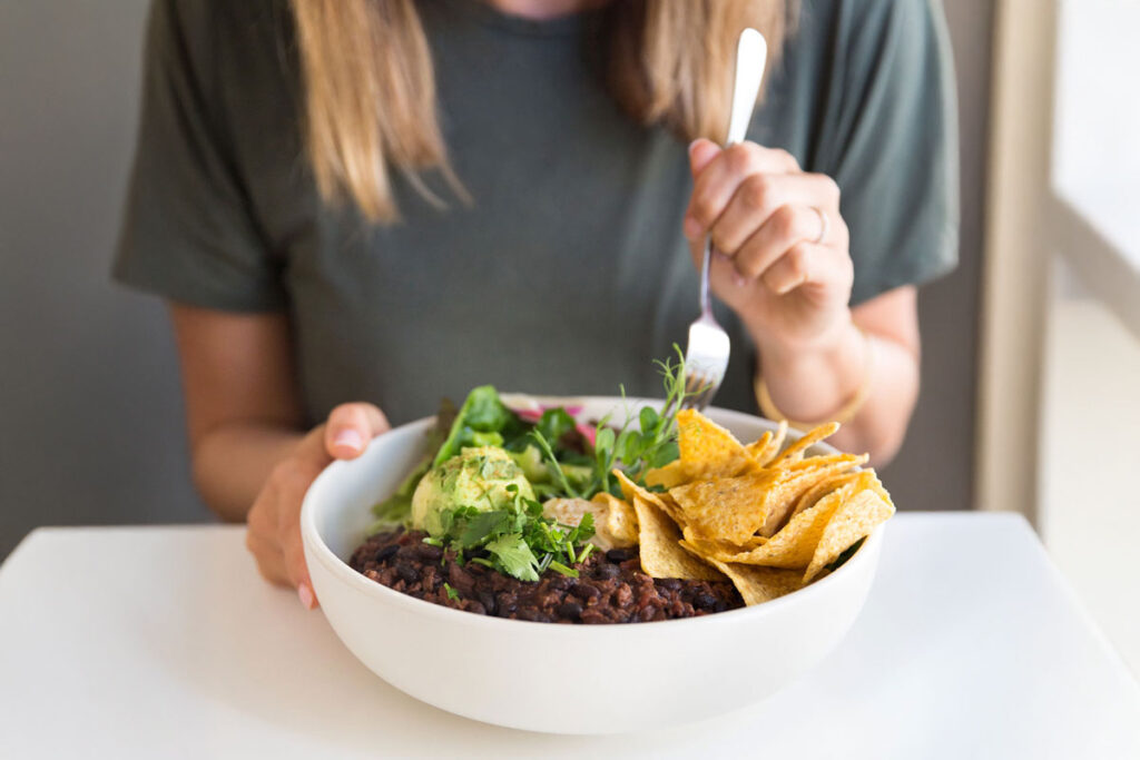 A person embraces a non-diet approach, savoring a vibrant bowl of black beans, avocado, fresh salad, and crisp tortilla chips, fork poised to enjoy every bite.